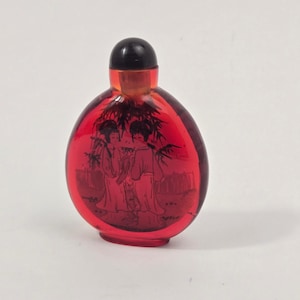 Puede incluir: Una botella de rapé de vidrio rojo translúcido con cuerpo redondeado y tapón negro. La superficie frontal presenta una detallada ilustración en tinta negra de dos figuras de pie bajo follaje de bambú, en un estilo artístico tradicional.