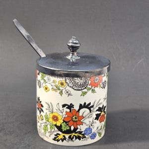 Puede incluir: Un tarro cilíndrico de cerámica para condimentos con base blanca y un colorido estampado floral en naranja, amarillo y azul. El tarro incluye una tapa de metal plateado pulido con un pomo central y una cuchara de metal a juego.