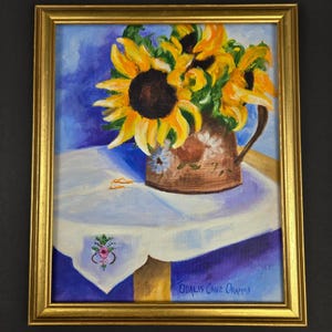 Puede incluir: Una pintura al óleo en un marco dorado que representa un ramo de girasoles de color amarillo brillante en una jarra de cerámica marrón. La jarra descansa sobre un paño blanco con un pequeño bordado floral, sobre un fondo azul. Firmado Odalis Cruz Oramas.