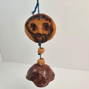 Puede incluir: Un adorno de arcilla colgante que presenta una cara circular con barba y rasgos faciales pintados en marrón oscuro, conectado por un hilo azul a dos pequeñas cuentas de arcilla y una pieza de arcilla esmaltada en marrón oscuro en forma de campana.