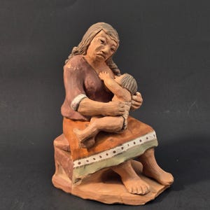 Op de afbeelding: Een handbeschilderd kleien beeldje van een zittend figuur die een baby voedt. De volwassene draagt een donker roodbruine top en een oranje rok met een lichtgroene zoom voorzien van een rij kleine bruine stippen. Het beeld heeft een matte afwerking.