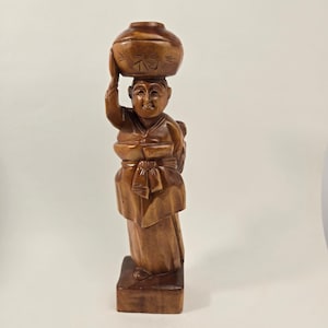 Puede incluir: Una figura de madera tallada a mano de una persona de pie sobre una base rectangular, con ropa tradicional y una faja en la cintura. La figura sostiene con ambas manos un recipiente grande y redondeado sobre la cabeza con marcas grabadas.