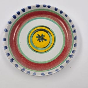 May include: A decorative ceramic plate with a white base, featuring concentric circles in red, green, and yellow. The center has a yellow circle with a black floral design. The rim is decorated with blue dots, creating a vibrant, handcrafted look.