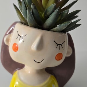 Puede incluir: Una maceta de cerámica caprichosa con forma de rostro de niña, con ojos cerrados y mejillas rosadas. La maceta tiene una parte superior amarilla con detalles triangulares blancos y está llena de una planta suculenta verde. Un artículo decorativo encantador.