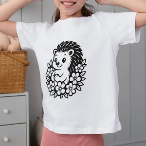 May include: White t-shirt with a black and white illustration of a hedgehog encircled by flowers. The hedgehog has a cheerful expression. The shirt is made of a soft material.