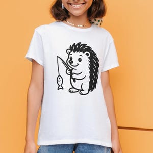 May include: White t-shirt with a black and white cartoon hedgehog fishing. The hedgehog is depicted with a fishing rod and a fish. The t-shirt has a round neck and short sleeves, a casual style.