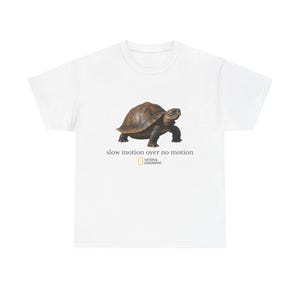 May include: White t-shirt featuring a realistic image of a tortoise with a brown shell and dark legs. The text "slow motion over no motion" is below the tortoise, with the National Geographic logo beneath the text.