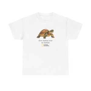 Puede incluir: Una camiseta blanca de manga corta con un gráfico de una tortuga marrón. Debajo de la tortuga, el texto "slow motion over no motion" aparece en negro, seguido del logotipo amarillo y blanco de National Geographic centrado en el pecho.