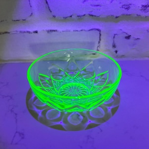 May include: A glowing green glass bowl with a starburst pattern. The bowl is round with a scalloped edge and sits on a surface that reflects its shape. The glass has a textured design.