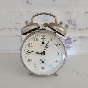 May include: A silver alarm clock with a white face and black hands. The clock has a second hand and a small dial for setting the alarm. The clock is set to 7:55. The clock has a JAZ logo on the face.