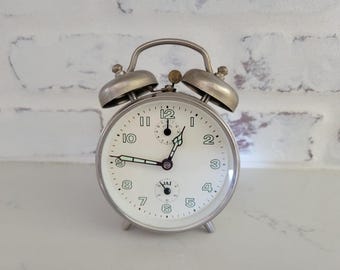 May include: A silver alarm clock with a white face and black hands. The clock has a second hand and a small dial for setting the alarm. The clock is set to 7:55. The clock has a JAZ logo on the face.