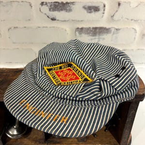May include: Vintage blue and white striped engineer's hat with a yellow and red patch that reads "THE RIO GRANDE GREAT SALT LAKE ROUTE RAILWAY". The word "ENGINEER" is embroidered in orange on the brim.