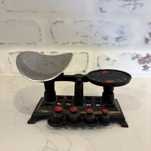 May include: A vintage black metal balance scale with a silver pan and a black platform with red floral accents. The scale includes a set of small weights with red tops, likely used for measuring ingredients or small items.