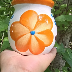 May include: A white ceramic flower pot with an orange rim and a large orange flower design. The flower has a blue center. The pot is held in front of a background of green leaves and tree branches.
