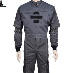May include: A grey jumpsuit with white stitching and black details. The top has a geometric design on the shoulders and chest, with two black rectangular patches. The long sleeves have black cuffs, and the trousers have reinforced knee patches. A costume.