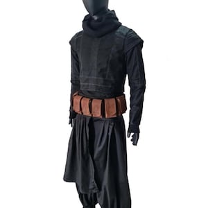 May include: A black long-sleeved tunic with a high cowl neck, paired with loose-fitting black trousers and a brown utility belt featuring multiple rectangular pouches. The outfit is displayed on a black mannequin against a white background.