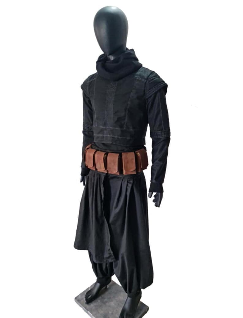 May include: A black long-sleeved tunic with a high cowl neck, paired with loose-fitting black trousers and a brown utility belt featuring multiple rectangular pouches. The outfit is displayed on a black mannequin against a white background.