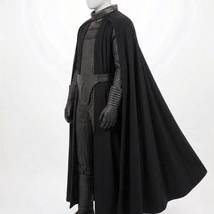 Op de afbeelding: Een volledig kostuum op een etalagepop, bestaande uit een lange zwarte cape, een donkerbruine hoge kraag, een gewatteerde zwarte jumpsuit, een donkerbruine utility-riem met een centrale ovale plaat, zwarte handschoenen en zwarte laarzen.