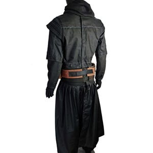 May include: A black costume ensemble featuring a long-sleeved jacket with structured shoulders and a high collar, paired with wide-leg trousers. A brown and black utility belt with rectangular panels is worn around the waist over the black clothing.
