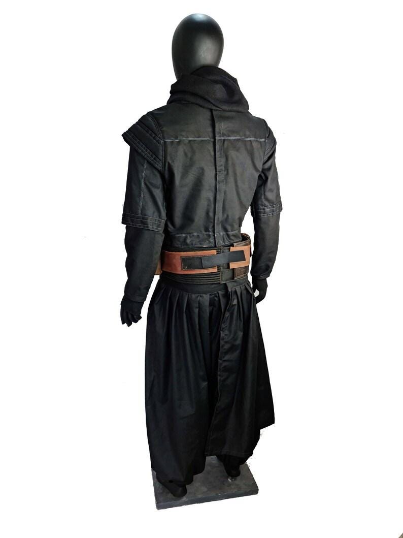 May include: A black costume ensemble featuring a long-sleeved jacket with structured shoulders and a high collar, paired with wide-leg trousers. A brown and black utility belt with rectangular panels is worn around the waist over the black clothing.
