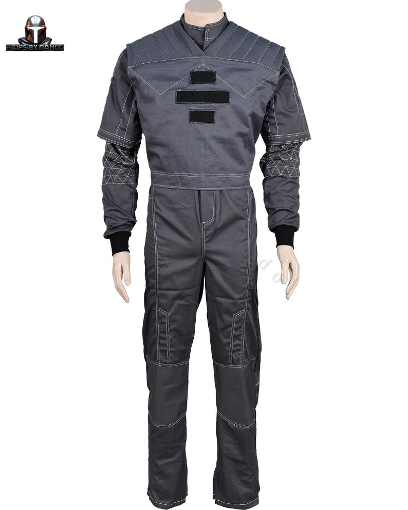 May include: A grey jumpsuit with white stitching and black details. The top has a geometric design on the shoulders and chest, with two black rectangular patches. The long sleeves have black cuffs, and the trousers have reinforced knee patches. A costume.