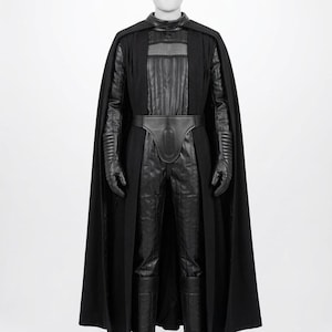 Op de afbeelding: Een zwart kostuum bestaande uit een lange cape, een textuurrijke jumpsuit met verticale stiksels, een brede riem met een ovaal detail in het midden, bijpassende handschoenen en hoge laarzen. De set bevat een borststuk met een hoge kraag.