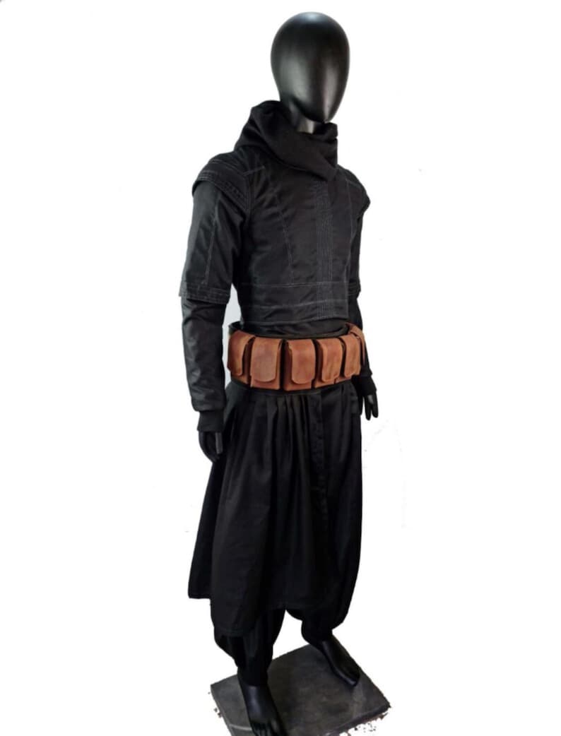 May include: A black hooded long-sleeved tunic with structured shoulder detailing and visible stitching, paired with loose-fitting black trousers. A tan leather utility belt featuring multiple rectangular pouches is worn around the waist.