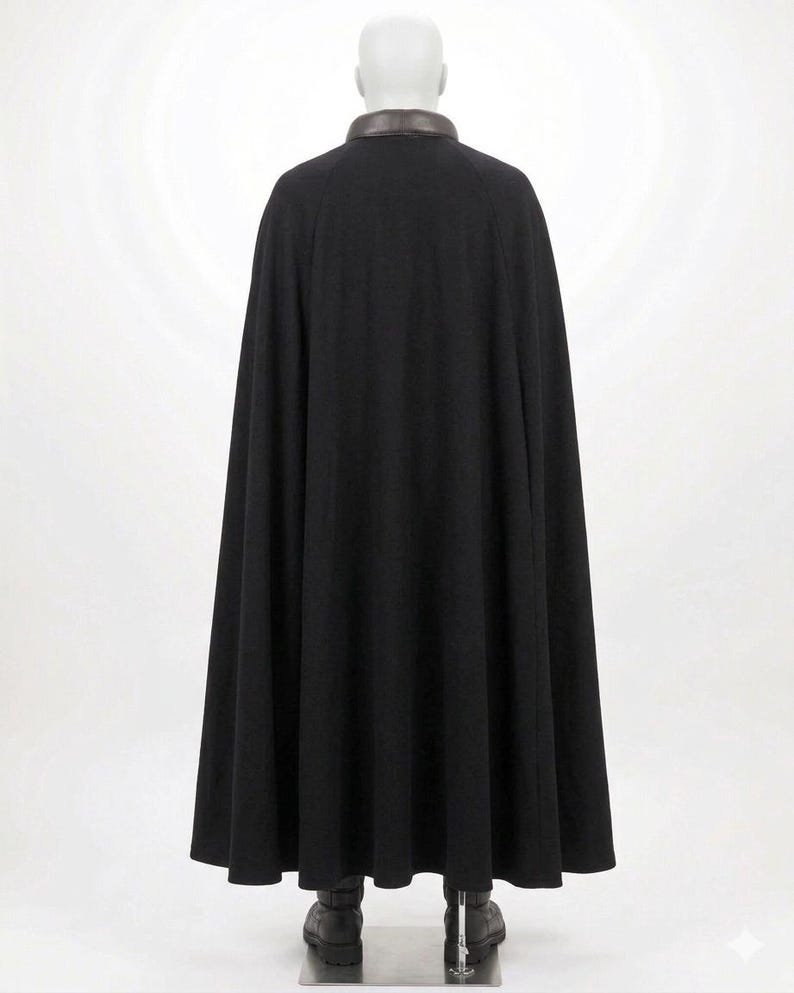 Op de afbeelding: Een zwarte cape tot op de grond met een gladde, donkerbruine kraag. Het kledingstuk wordt getoond op een etalagepop en heeft een volledig, vloeiend silhouet dat vanaf de schouders tot aan de zoom valt en de rug van de figuur bedekt.