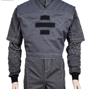 Puede incluir: Mono gris con costuras blancas y detalles negros. La parte superior presenta un dise&ntilde;o de cuello en V, hombros acolchados y tres parches rectangulares negros. Las mangas tienen patrones geom&eacute;tricos y terminan con pu&ntilde;os negros. Los pantalones tienen una costura blanca en la parte delantera.