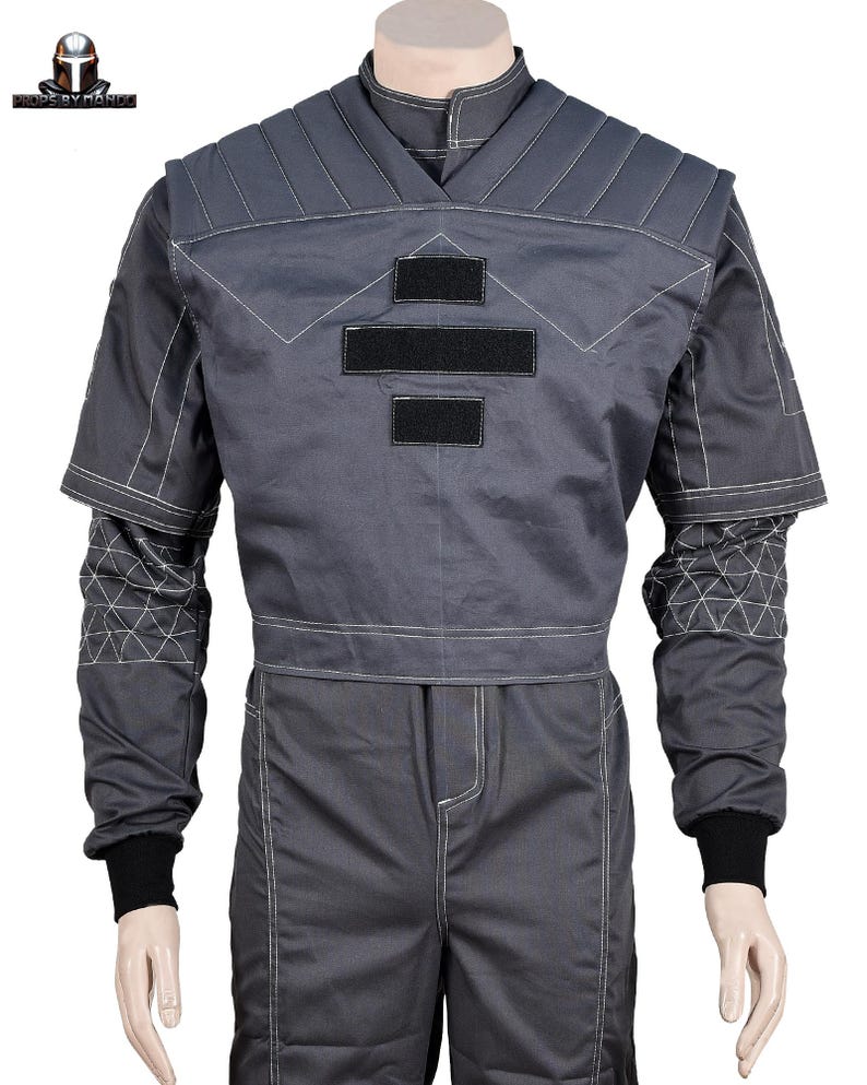 Puede incluir: Mono gris con costuras blancas y detalles negros. La parte superior presenta un dise&ntilde;o de cuello en V, hombros acolchados y tres parches rectangulares negros. Las mangas tienen patrones geom&eacute;tricos y terminan con pu&ntilde;os negros. Los pantalones tienen una costura blanca en la parte delantera.