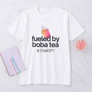 Puede incluir: Camiseta blanca con el texto "fueled by boba tea & ChatGPT" y un gráfico de una bebida de té boba rosa y naranja. Una bebida rosa con una pajita es visible en el fondo.