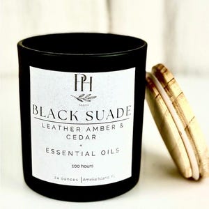 Può includere: Un barattolo di candela nero con un'etichetta bianca con la scritta "BLACK SUADE LEATHER AMBER & CEDAR + ESSENTIAL OILS". La candela ha un coperchio di legno e pesa 680 grammi.