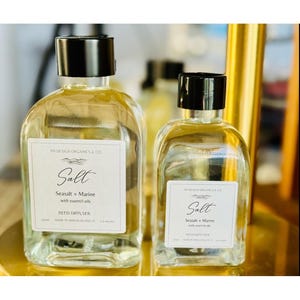 May include: Two clear glass reed diffuser bottles with black lids. The bottles have white labels with the word "Salt" and "Seasalt + Marine" printed on them. The bottles are filled with a light yellow liquid.