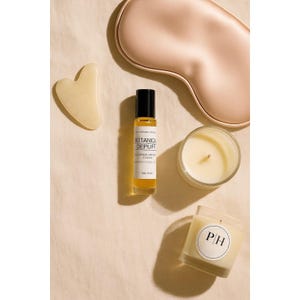 Op de afbeelding: Een flatlay met zelfzorgartikelen. Het bevat een lichtroze oogmasker, een gele gua sha tool, een rollerfles met de tekst "Botanica Depuff" en twee kaarsen in glazen potten. Eén kaars heeft de initialen "PH" op een label.