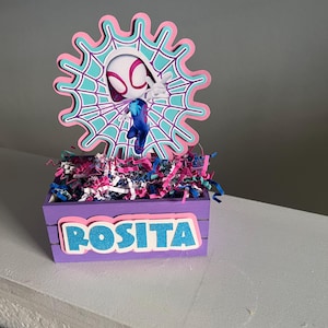 Può includere: Una scatola di legno viola con il nome "ROSITA" in lettere blu glitterate. La scatola è riempita di carta sminuzzata colorata e sormontata da un ritaglio del personaggio di Spider-Gwen in rosa, turchese e bianco.