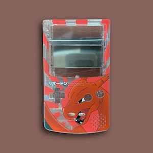 May include: Clear plastic Game Boy shell featuring a red and orange Charizard design with Japanese text. The design includes a rising sun motif and the Nintendo logo. The shell has a transparent screen area and button details.