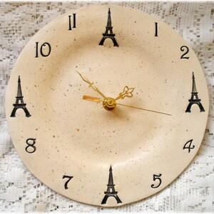 May include: A cream-colored clock with black numbers and Eiffel Tower designs. The clock hands are gold.