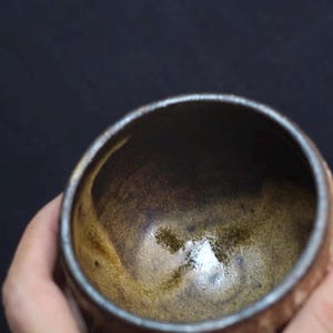 May include: A handmade ceramic bowl with a dark brown interior and a lighter brown exterior. The bowl has a smooth, glossy finish and a slightly textured surface. The rim of the bowl is a darker shade of brown. The bowl is held in a hand.