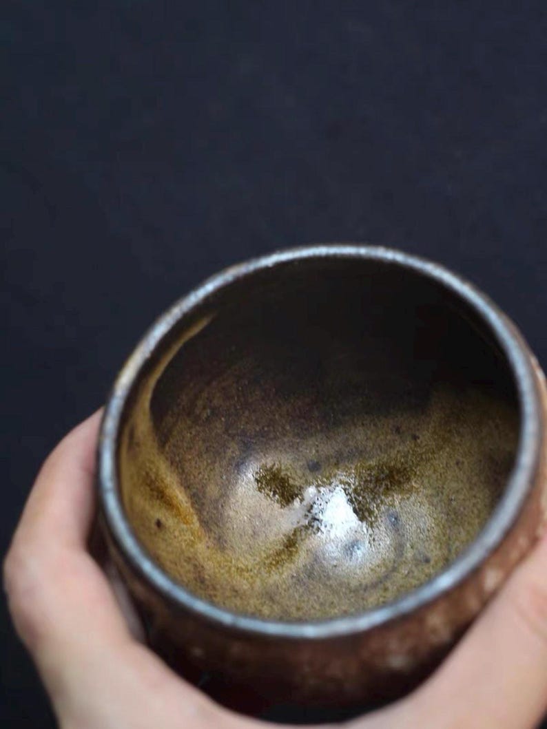 May include: A handmade ceramic bowl with a dark brown interior and a lighter brown exterior. The bowl has a smooth, glossy finish and a slightly textured surface. The rim of the bowl is a darker shade of brown. The bowl is held in a hand.