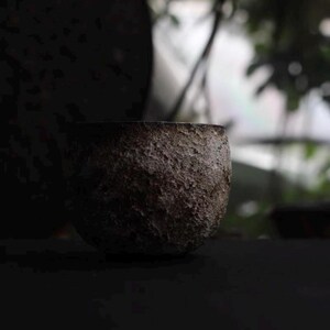 May include: A small, textured, dark brown ceramic bowl. The bowl has a rough, uneven surface and a rounded shape. The background is blurred, with hints of green foliage and a dark, circular object. The lighting is dim, creating a moody atmosphere.