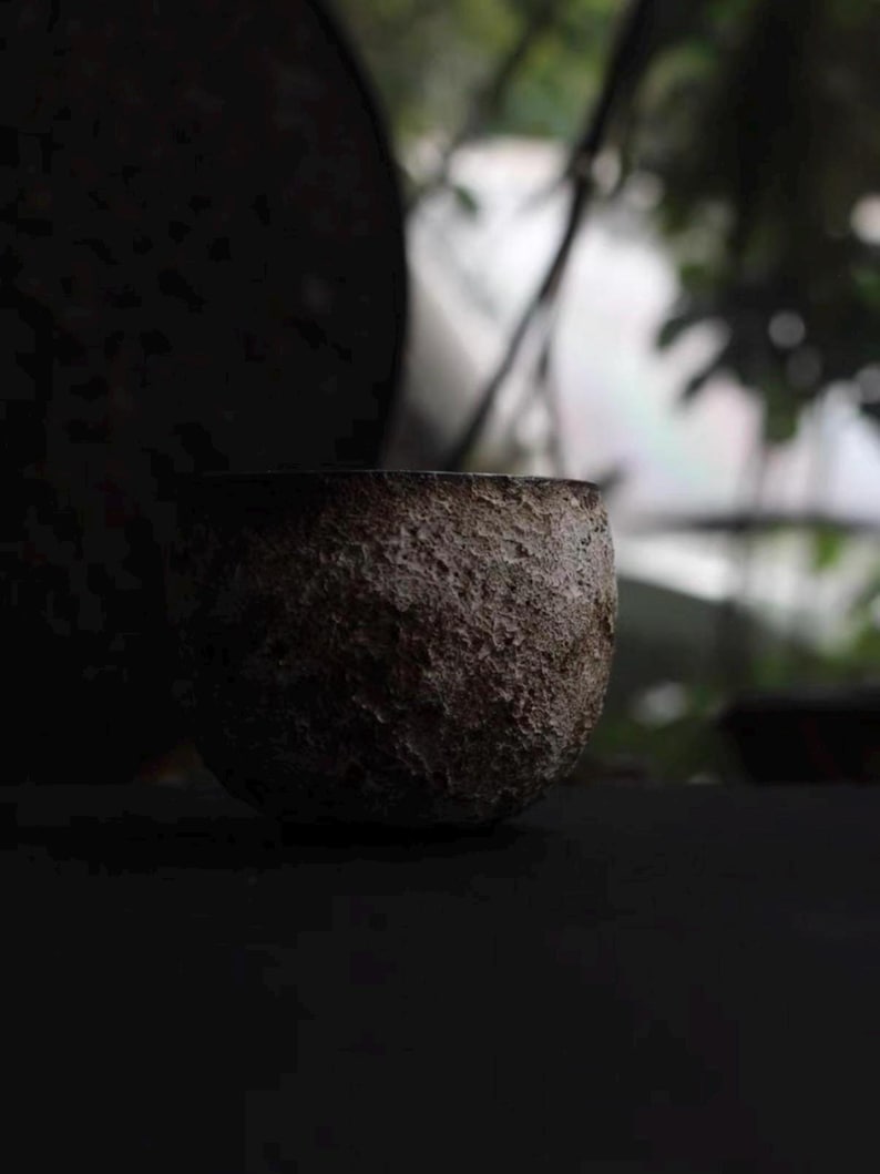May include: A small, textured, dark brown ceramic bowl. The bowl has a rough, uneven surface and a rounded shape. The background is blurred, with hints of green foliage and a dark, circular object. The lighting is dim, creating a moody atmosphere.