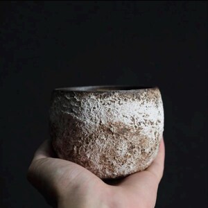 May include: A handcrafted, textured ceramic bowl with a rough, white-washed exterior over a brown base. The bowl has a rounded shape and a dark interior, perfect for serving or display.