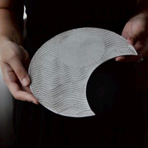Op de afbeelding: Een halvemaanvormige, grijze keramische plaat met een textuur van golven en een glad, cirkelvormig midden. De plaat wordt vastgehouden, waardoor het unieke ontwerp tegen een donkere achtergrond zichtbaar is.