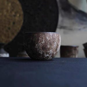 May include: A small, round, textured ceramic bowl with a rough, earthy appearance. The bowl is a muted brown color with lighter patches, suggesting a natural, handcrafted aesthetic. The background is dark, emphasizing the bowl's form.