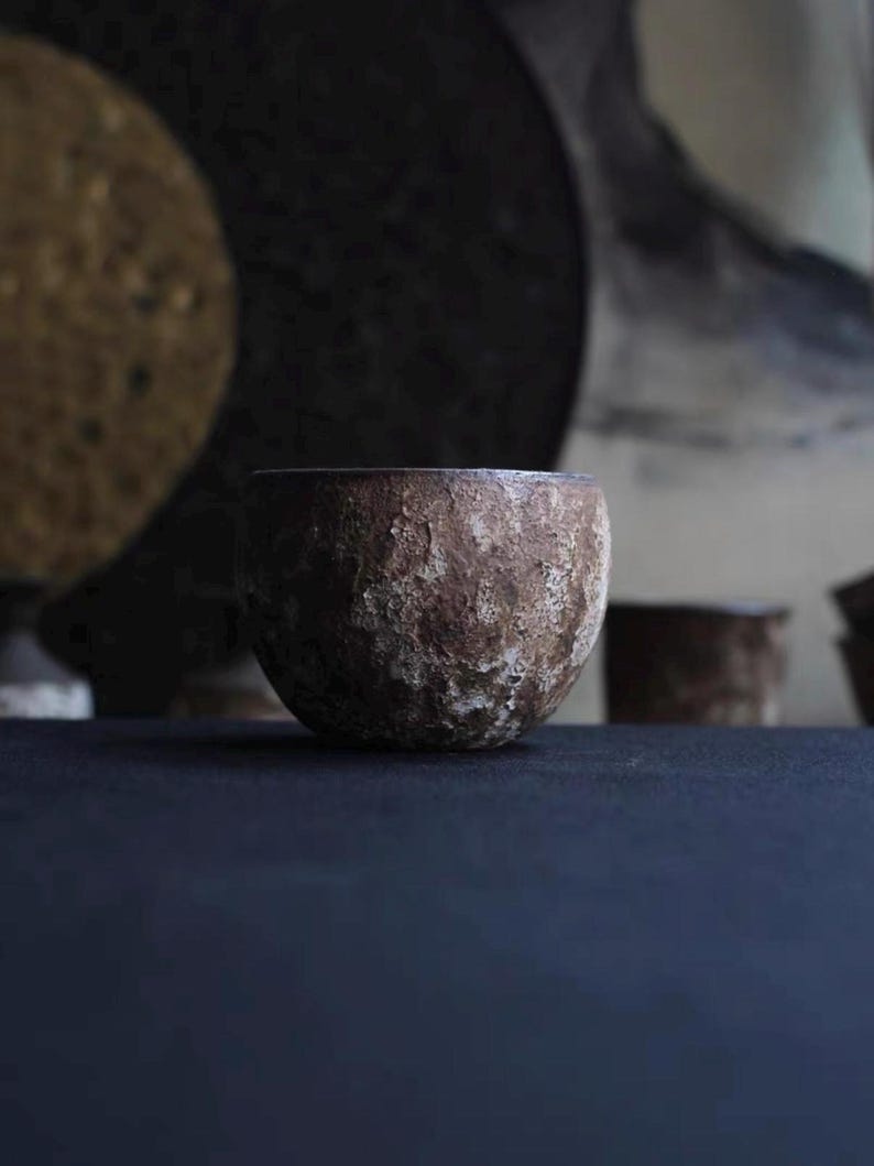 May include: A small, round, textured ceramic bowl with a rough, earthy appearance. The bowl is a muted brown color with lighter patches, suggesting a natural, handcrafted aesthetic. The background is dark, emphasizing the bowl's form.