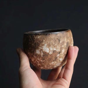 May include: A small, handcrafted ceramic bowl with a textured, brown exterior and a dark interior. The bowl is held in a hand against a dark background, showcasing its rustic aesthetic. The bowl's shape is round and the rim is smooth.
