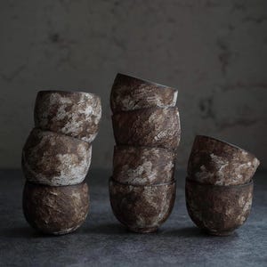 May include: A collection of handmade ceramic bowls in varying stacks. The bowls have a textured, earthy appearance with a mottled brown and white glaze. The bowls are arranged on a dark gray surface, creating a rustic and minimalist aesthetic.