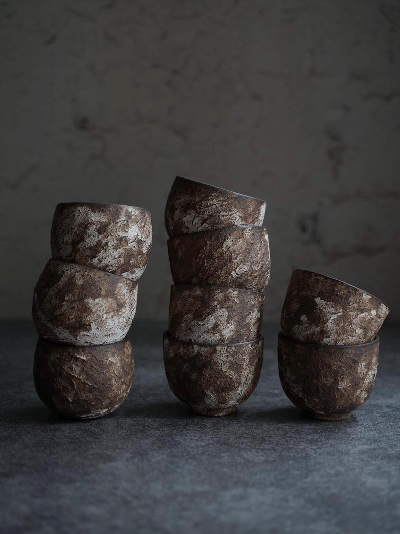 May include: A collection of handmade ceramic bowls in varying stacks. The bowls have a textured, earthy appearance with a mottled brown and white glaze. The bowls are arranged on a dark gray surface, creating a rustic and minimalist aesthetic.