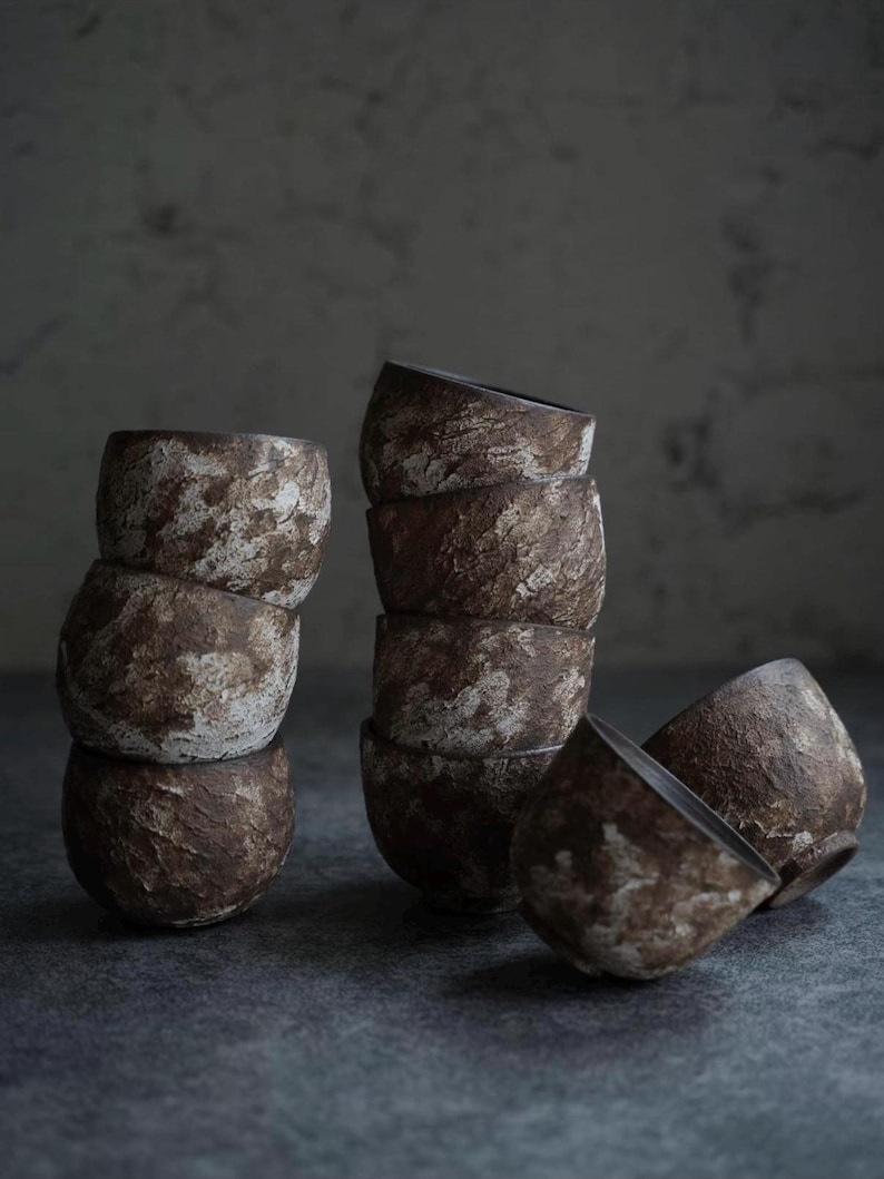 May include: A collection of handmade ceramic bowls in a rustic style. The bowls are stacked in groups of three and four, with a few scattered nearby. The textured surface has a brown and white mottled appearance, giving them a natural, earthy look.