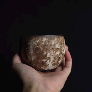 May include: A small, round, ceramic bowl with a textured, brown surface and white accents. The bowl is held in a hand against a dark background. The bowl's shape is simple and organic, suggesting a handmade quality.